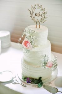 A white wedding cake with flowers and a wooden topper.